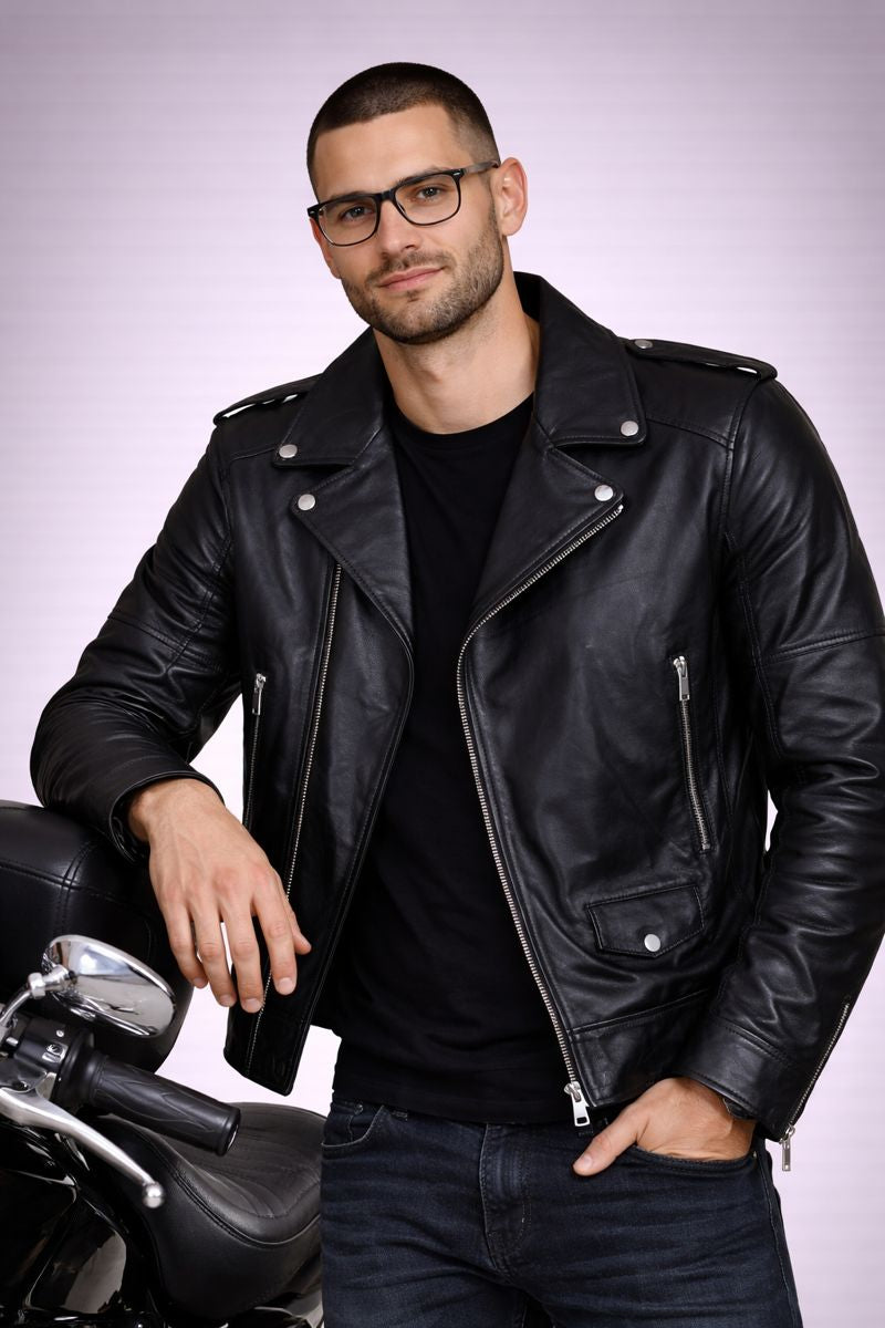 Man wearing a black leather jacket and glasses, standing next to a motorcycle on a light gray background