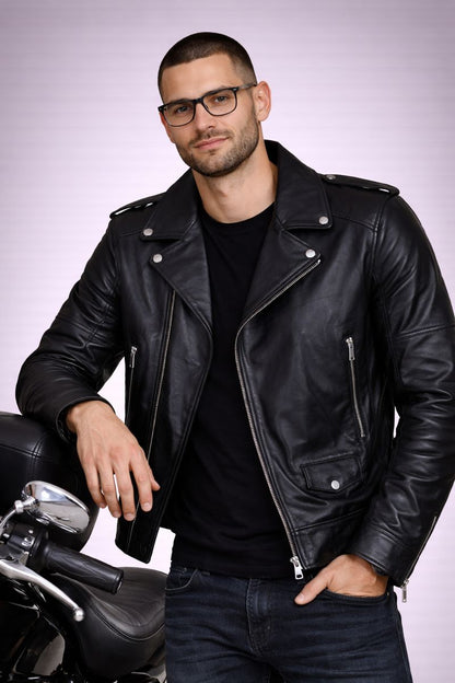 Man wearing a black leather jacket and glasses, standing next to a motorcycle on a light gray background